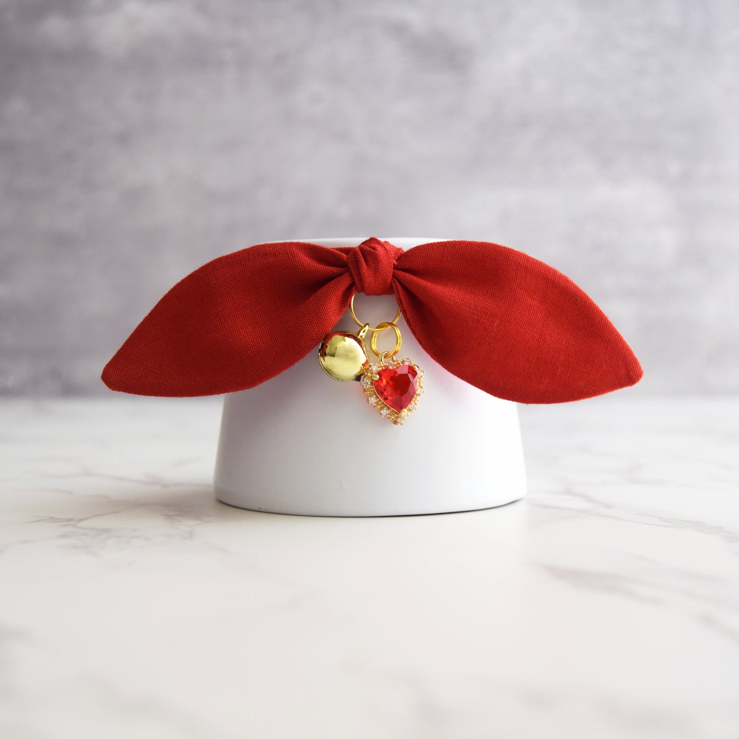 A red velvet bow cat collar with dangling gold bell and a heart shaped red gemstone, displayed on a white cylindrical stand against a gray background.