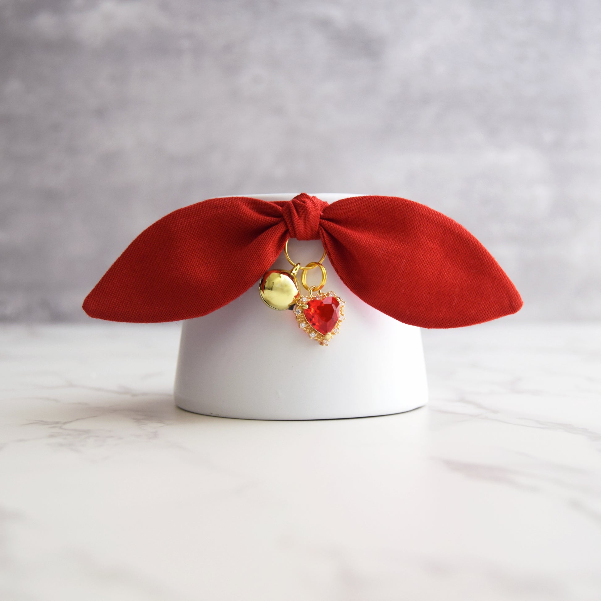 A red velvet bow cat collar with dangling gold bell and a heart shaped red gemstone, displayed on a white cylindrical stand against a gray background.