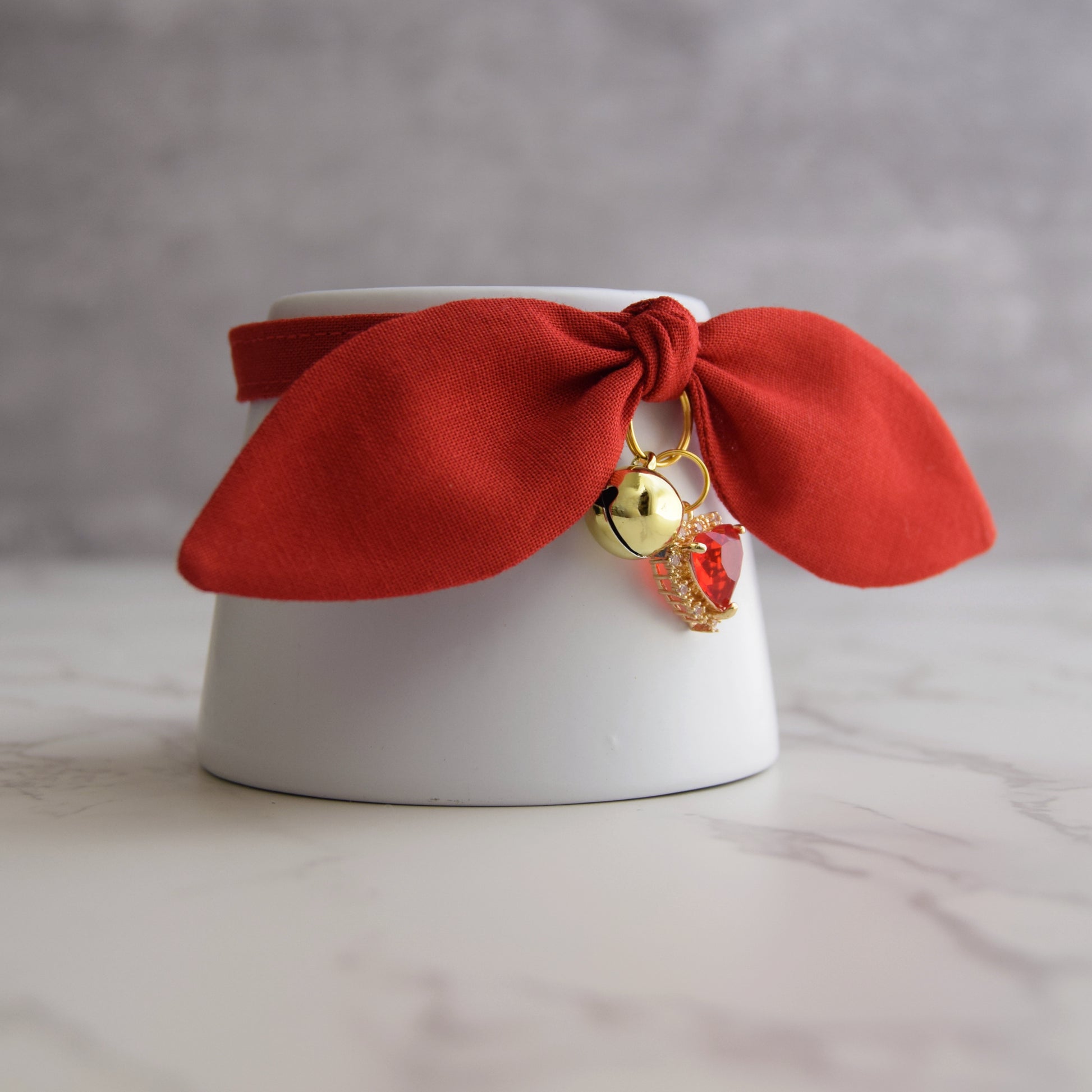 A red fabric pet collar with floppy ears and small dangling charms, including a gold bell and a red crystal charm, displayed on a white ceramic cup against a soft gray background.