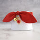 A red fabric bow collar with gold-tone hardware, featuring a small gold bell and a red heart-shaped charm with gold detailing, displayed on a white cylindrical stand.