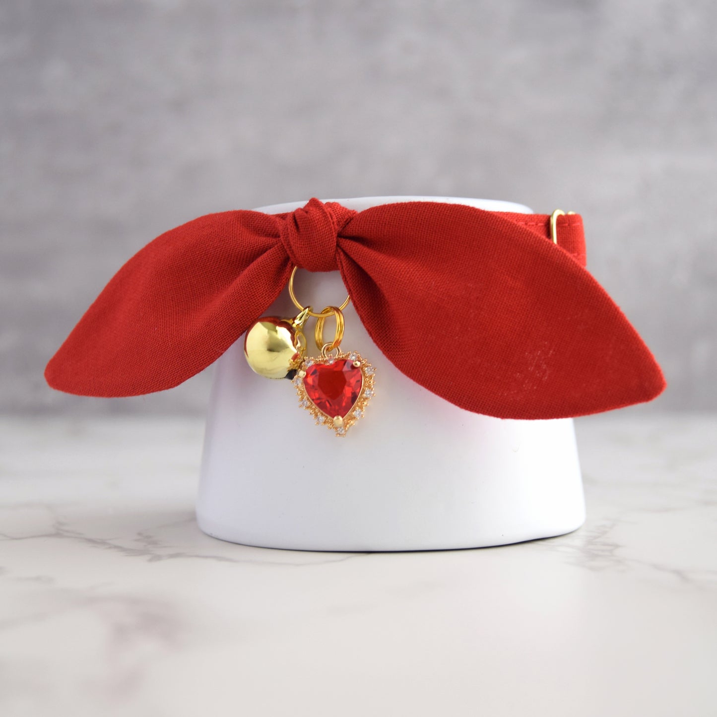 A red fabric bow collar with gold-tone hardware, featuring a small gold bell and a red heart-shaped charm with gold detailing, displayed on a white cylindrical stand.