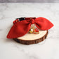 A red fabric pet collar with a large bow, gold bell, and red heart charm, displayed on a rustic wood slice coaster over a white marble surface.