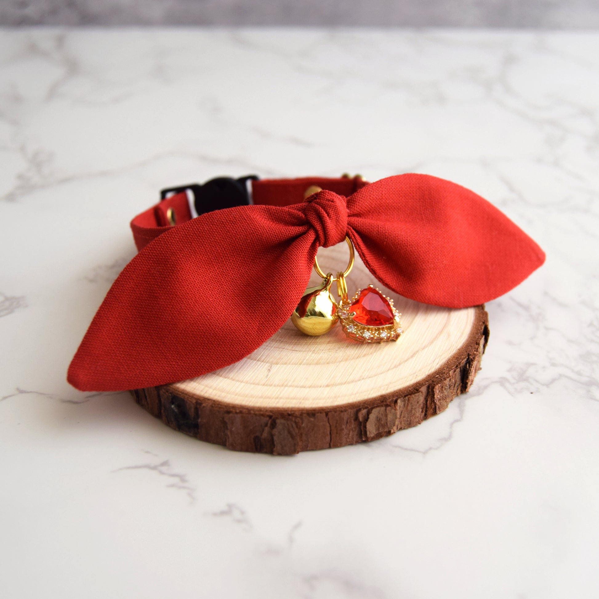 A red fabric pet collar with a large bow, gold bell, and red heart charm, displayed on a rustic wood slice coaster over a white marble surface.
