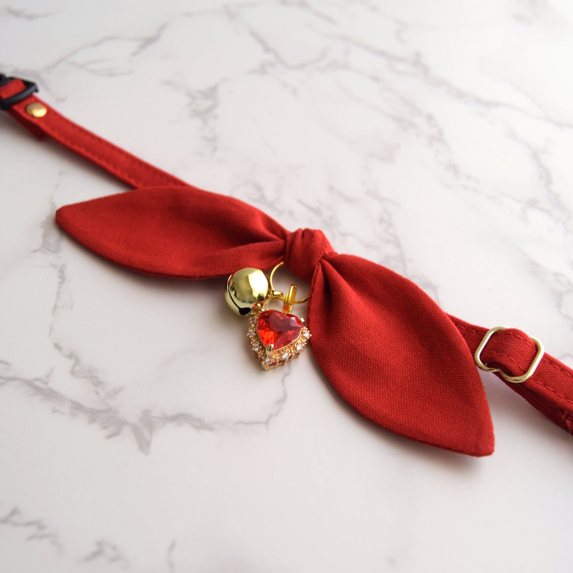 A red fabric pet collar with a bow detail, gold-tone metal hardware, and a dangling heart-shaped crystal charm, displayed on a white marble surface.
