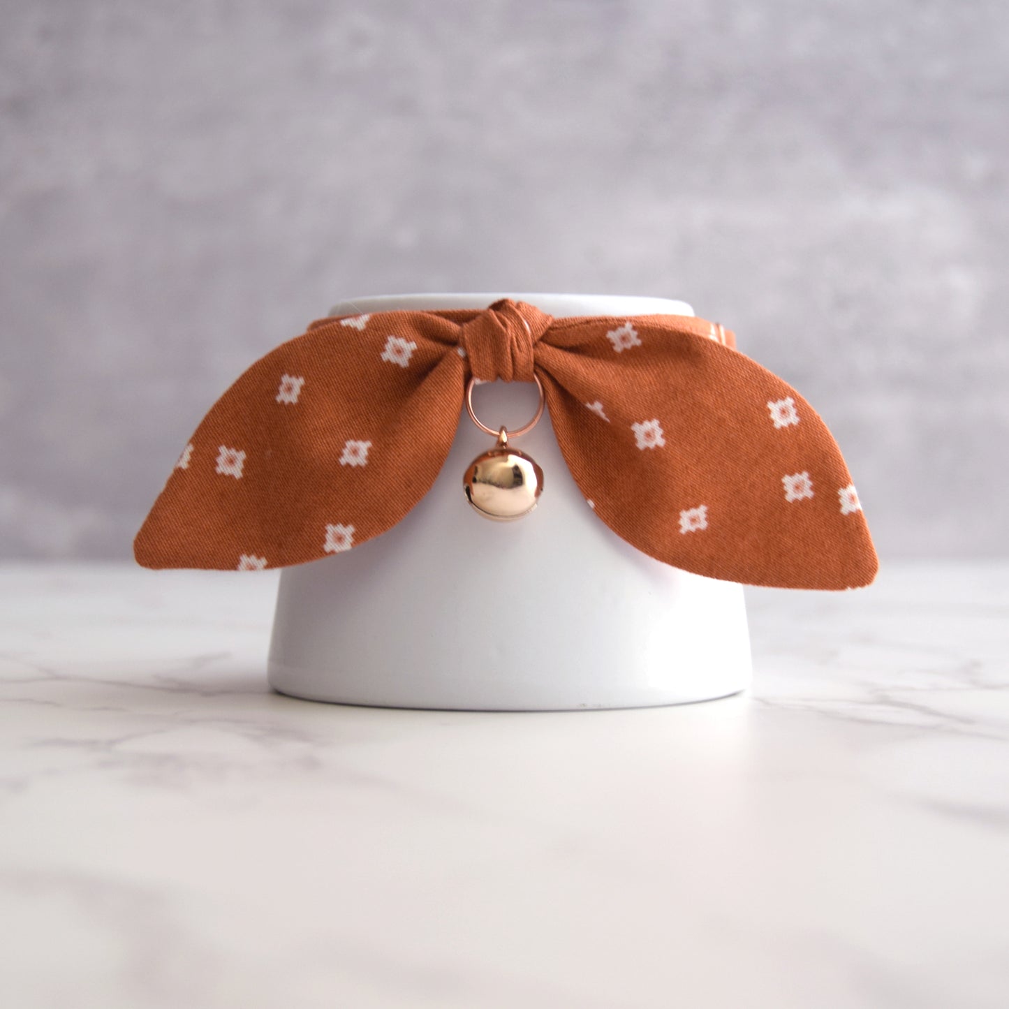 A white ceramic cup or small vase with a rust-orange fabric bow cat collar tied around the rim, decorated with small white geometric motifs and a dangling metallic gold bell charm, set on a light marble surface against a soft gray background.