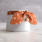 An orange-brown fabric pet collar with a white geometric pattern, tied in a bow around a white ceramic cup, and featuring a small rose gold bell.
