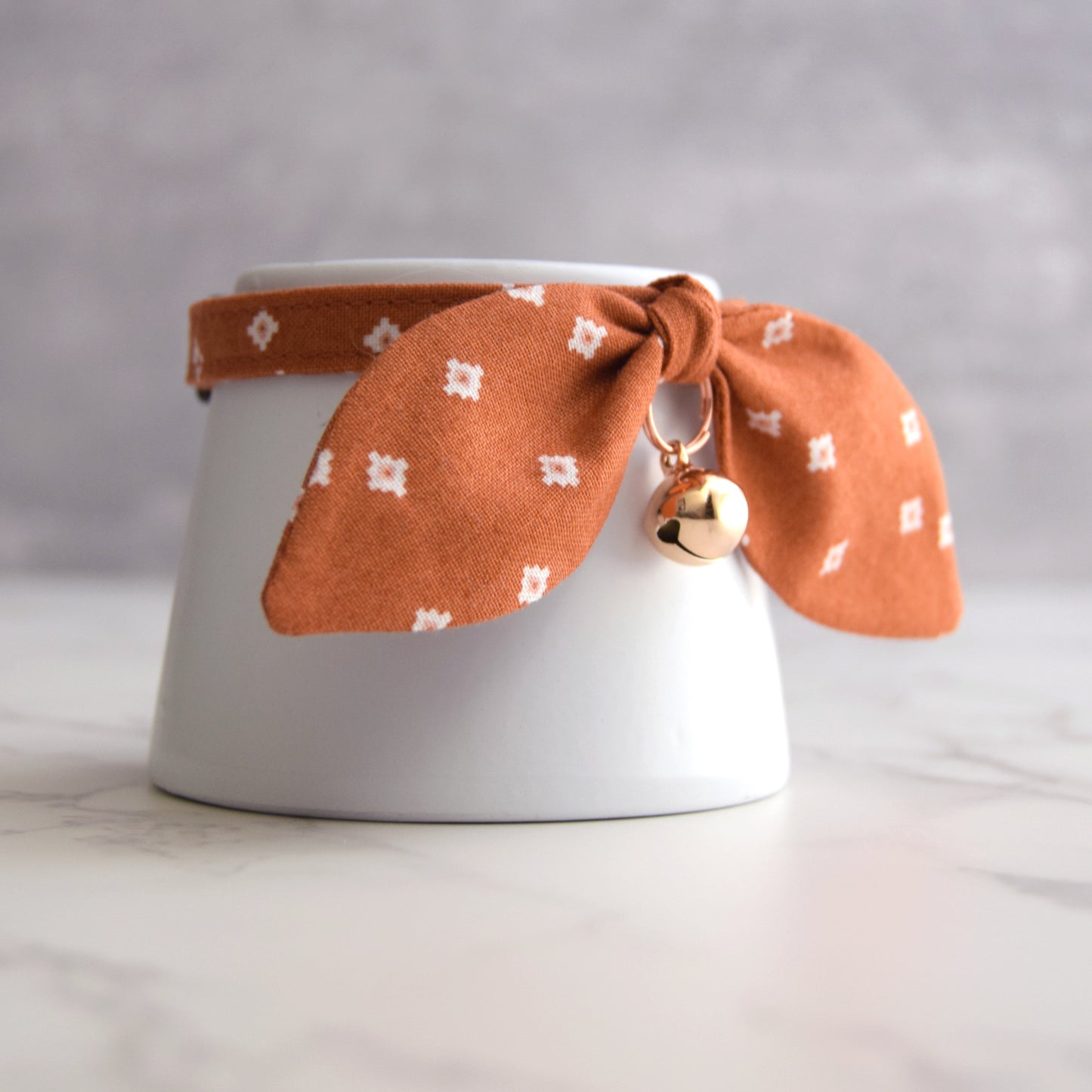 An orange-brown fabric pet collar with a white geometric pattern, tied in a bow around a white ceramic cup, and featuring a small rose gold bell.