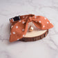 A terracotta orange fabric pet collar with a large bow, small white geometric pattern, and a rose gold bell, displayed on a rustic wood slice over a white marble background.