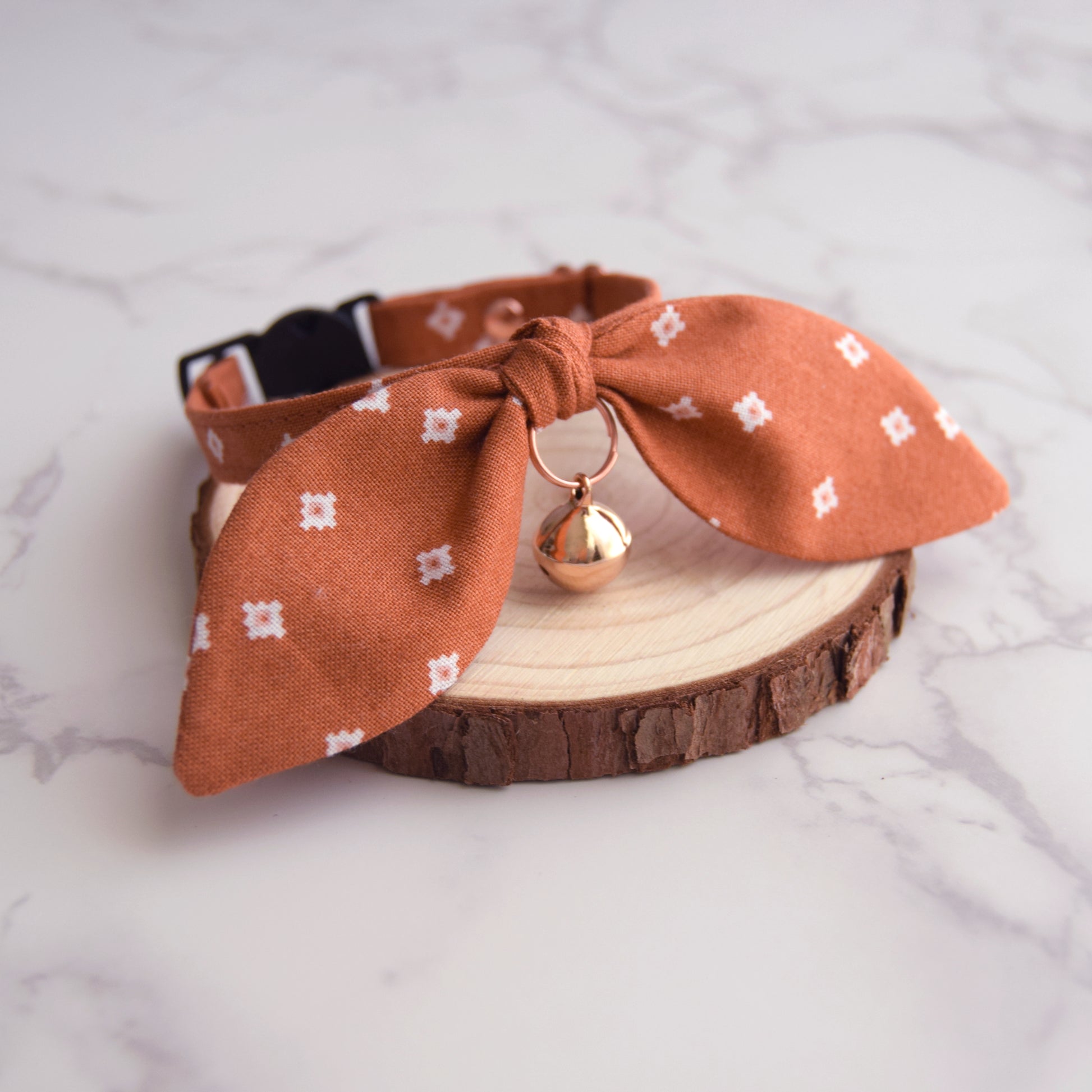 A terracotta orange fabric pet collar with a large bow, small white geometric pattern, and a rose gold bell, displayed on a rustic wood slice over a white marble background.