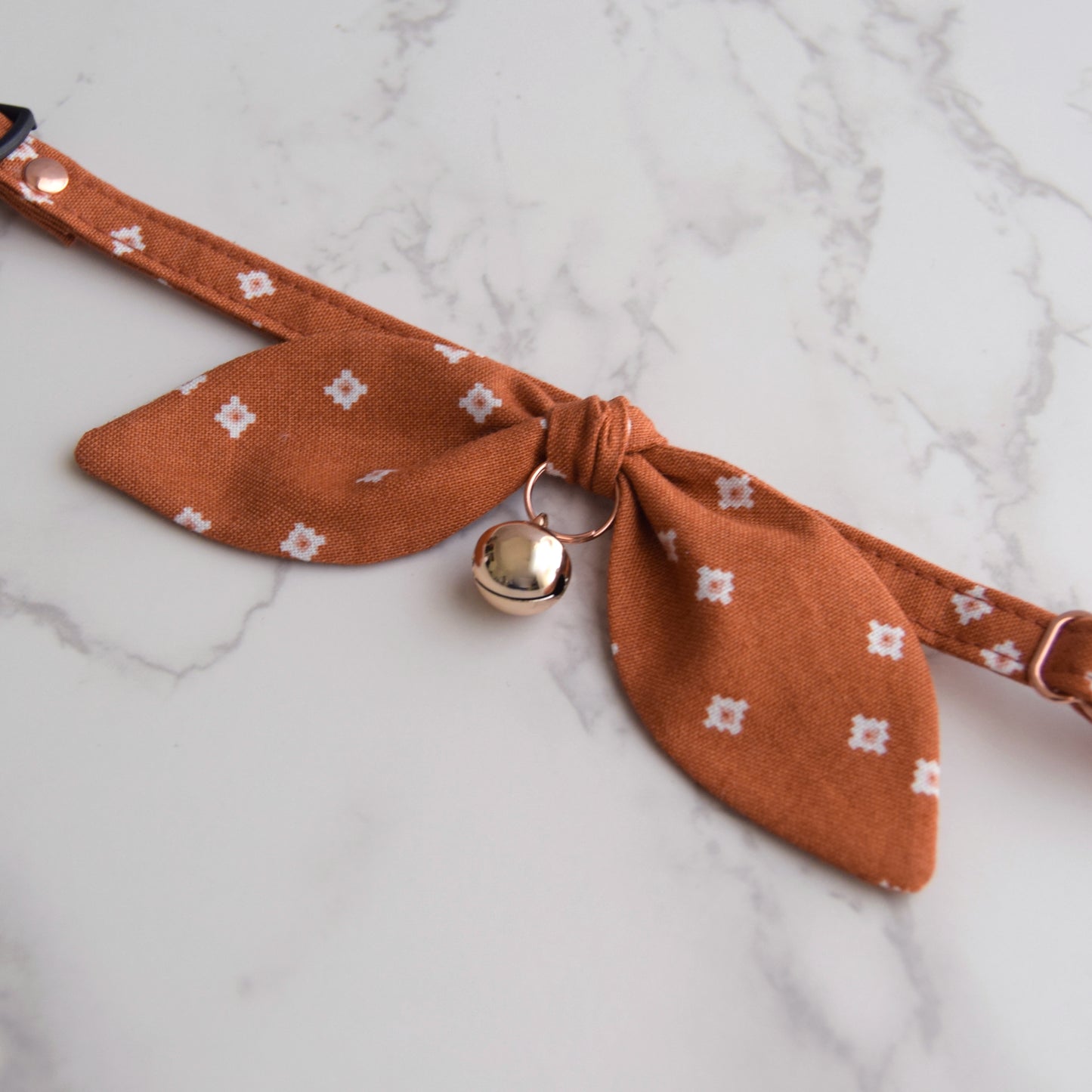 A terracotta orange fabric pet collar with a small white patterned bow and a rose gold bell, displayed on a white marble surface.