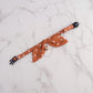 An orange-brown pet collar with a matching fabric bow tie, small rose gold bell, and black plastic hardware, laid flat on a white marble background.