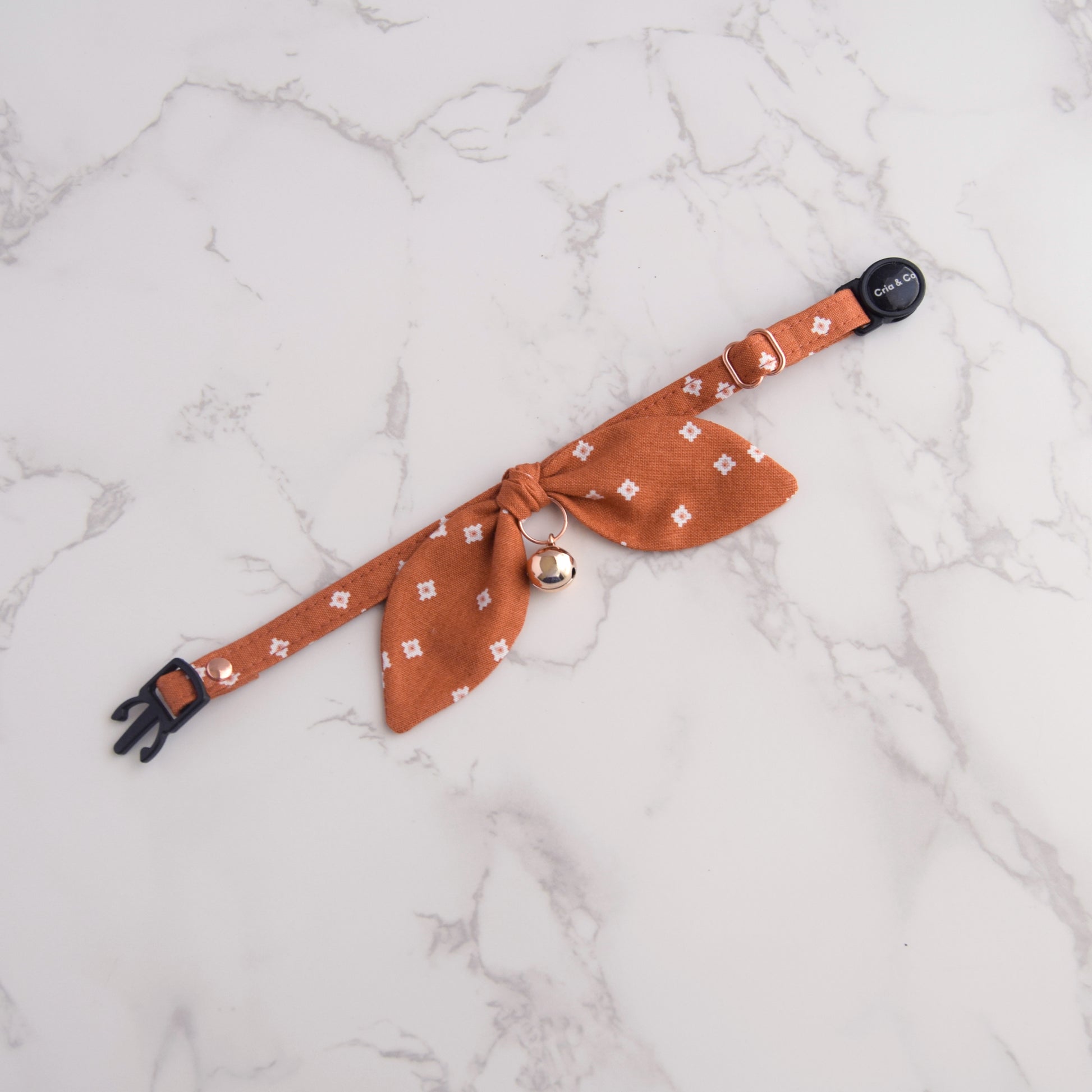 An orange-brown pet collar with a matching fabric bow tie, small rose gold bell, and black plastic hardware, laid flat on a white marble background.
