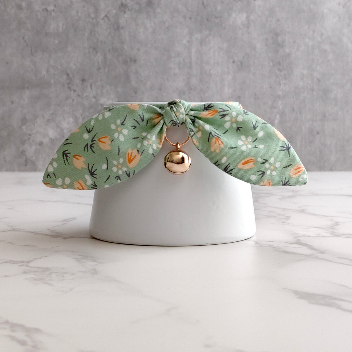 A sage green fabric pet collar with a white and orange floral pattern, tied in a bow on a white ceramic stand with a small gold bell.