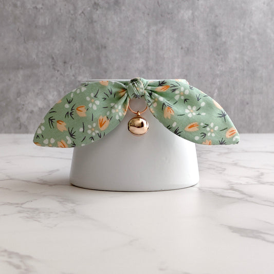 A sage green fabric pet collar with a white and orange floral pattern, tied in a bow on a white ceramic stand with a small gold bell.