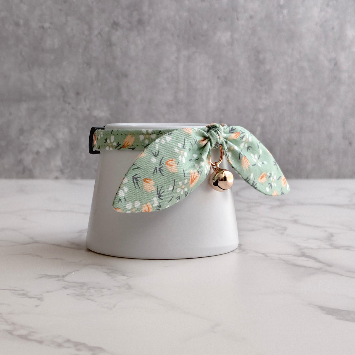 A white ceramic cup or small planter with a green floral fabric band around the rim and a small gold bell hanging from the tie, set on a light marble surface against a gray textured background.