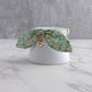 A green floral fabric pet collar with a small gold bell, displayed on a white ceramic stand over a white marble surface against a gray textured background.