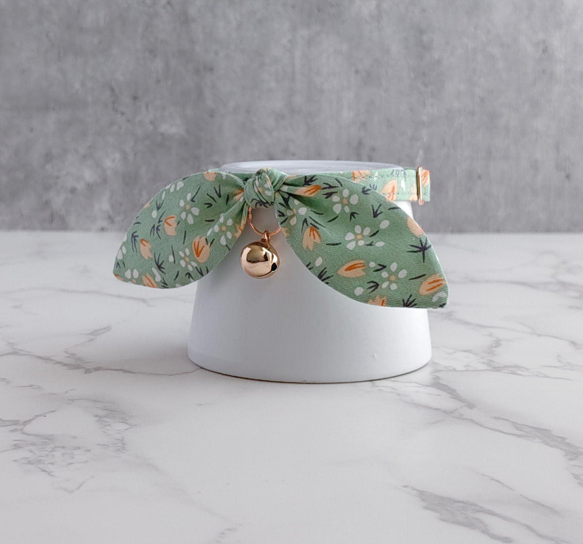 A green floral fabric pet collar with a small gold bell, displayed on a white ceramic stand over a white marble surface against a gray textured background.