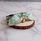 A pet collar with a large bow made from sage green fabric printed with orange carrots and white flowers, featuring a small gold bell, displayed on a round wood slice slice on a white marble surface.