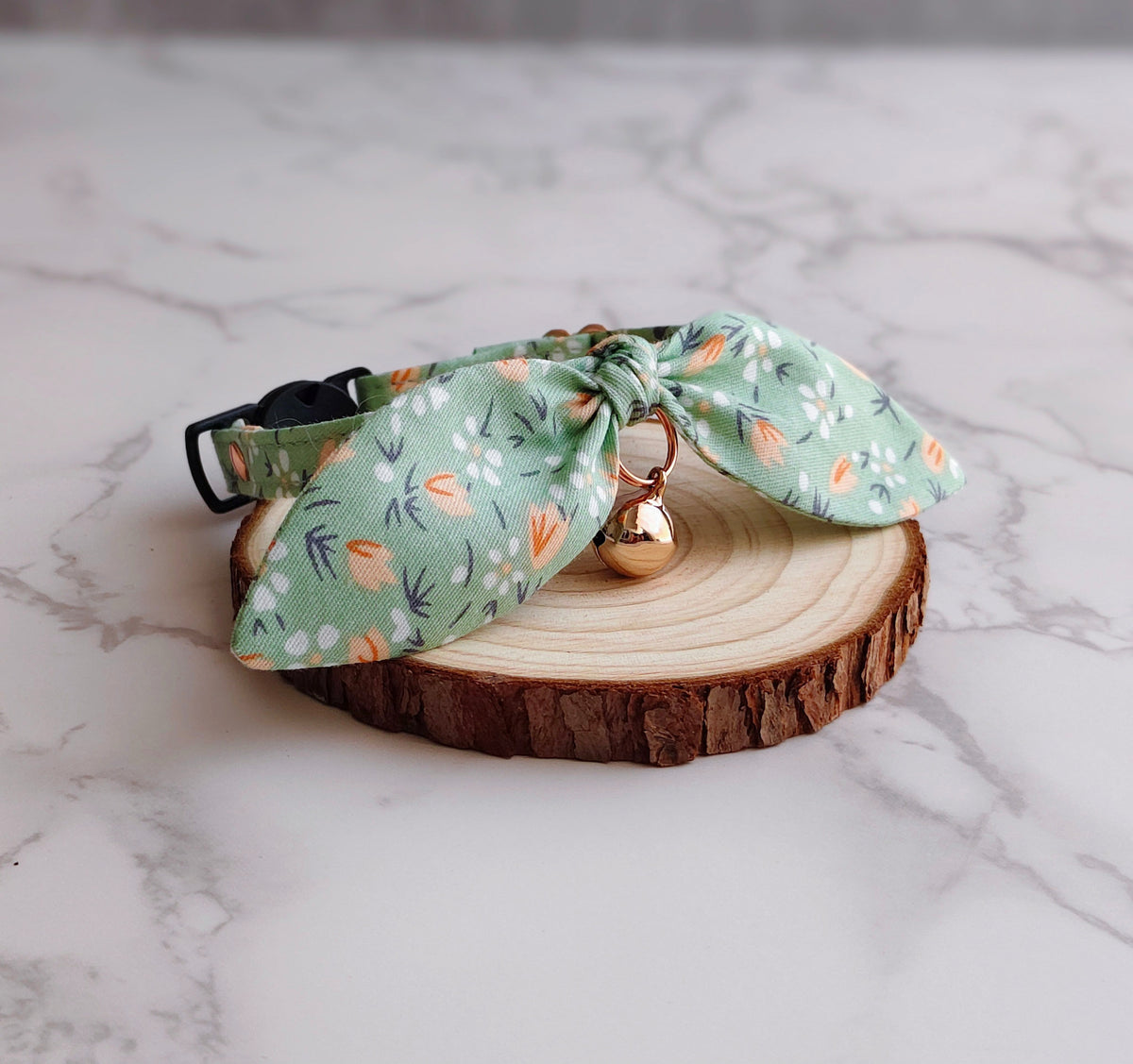 A pet collar with a large bow made from sage green fabric printed with orange carrots and white flowers, featuring a small gold bell, displayed on a round wood slice slice on a white marble surface.