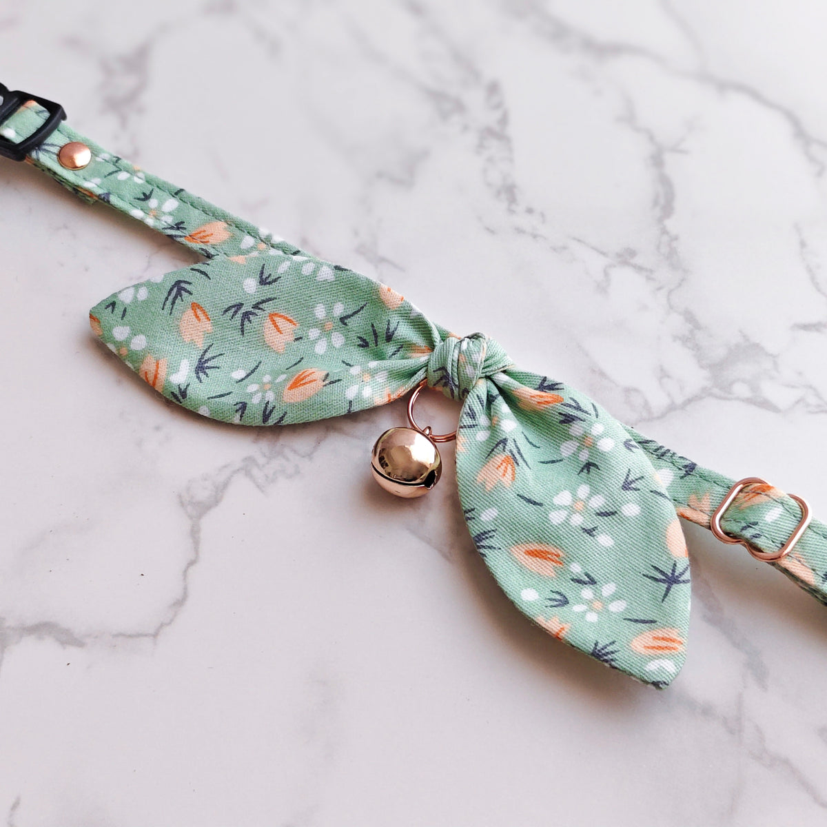 A mint green fabric pet collar with a floral print, featuring a central bow, a small gold bell, and adjustable metal hardware, laid flat on a white marble background.